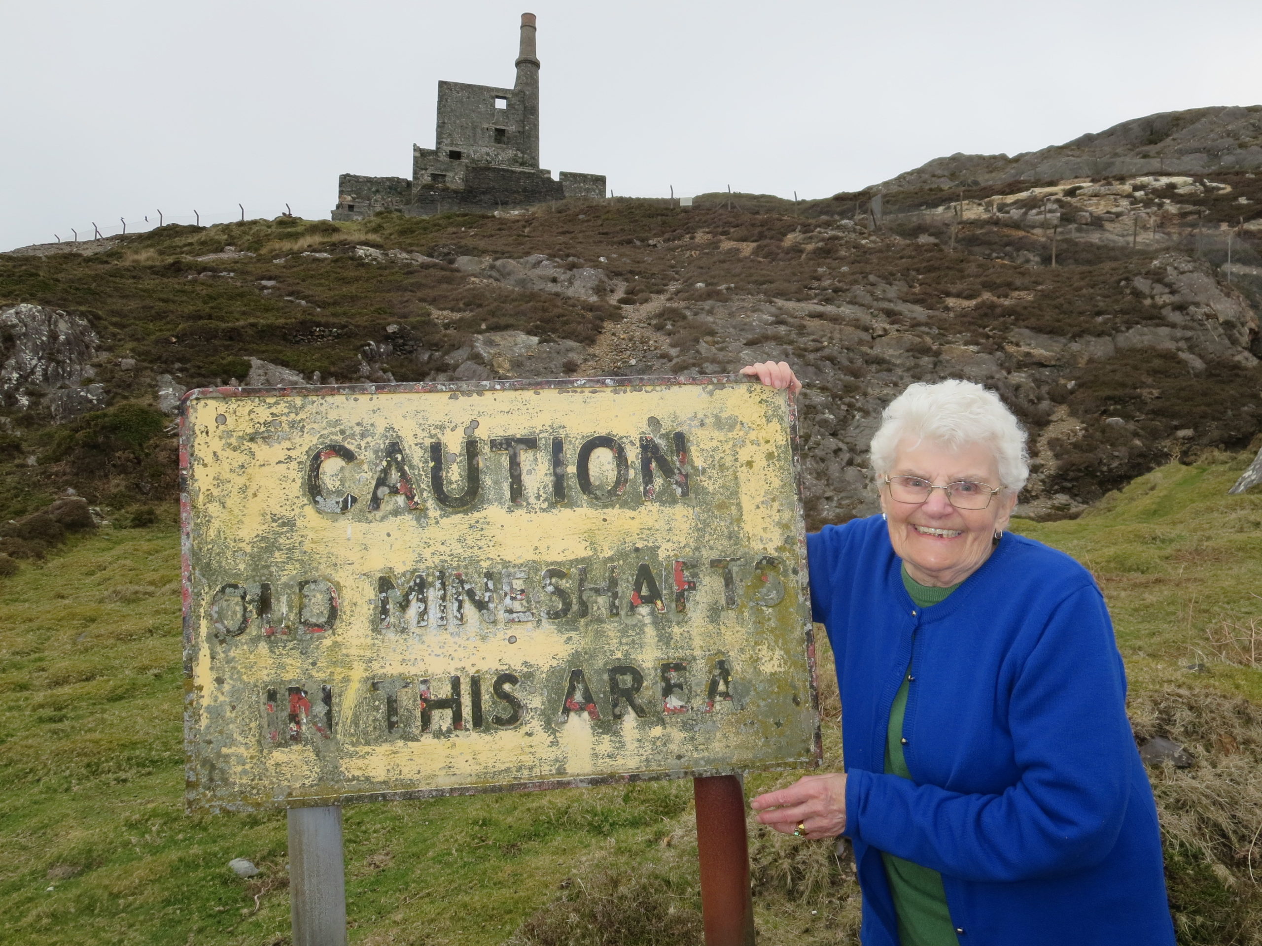 DP Lindegaard next to a sign saying "Caution Old Mineshafts in this area" with Allihies Mineworks in thr background