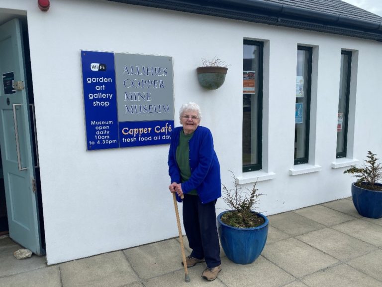 DP Lindegaard outside the Allihies mIning Museum. She is wearing a blue cardigan and leaning on a walking stick. The white walls of the musiem are behind her.